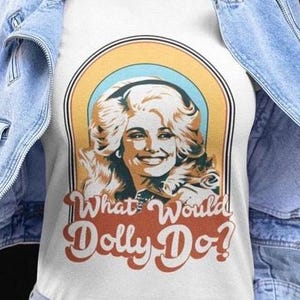 Puede incluir: Una camiseta blanca con un estampado gráfico de una mujer rubia con cabello voluminoso dentro de un arco de estilo retro. El texto "What Would Dolly Do?" está impreso en una fuente naranja ondulada y negrita debajo de la ilustración, bajo una chaqueta vaquera.
