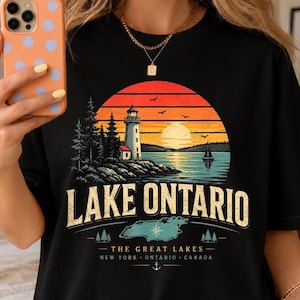 Peut inclure: Un t-shirt noir avec une impression graphique représentant un phare sur une rive rocheuse au bord d'un lac au coucher du soleil. Le texte indique LAKE ONTARIO, THE GREAT LAKES, NEW YORK, ONTARIO, CANADA. Le motif inclut une carte, une rose des vents et une ancre.
