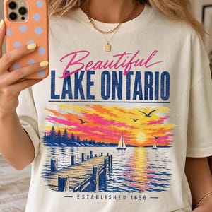 Peut inclure: Un t-shirt blanc cassé avec un motif représentant un coucher de soleil sur le lac Ontario, incluant un ponton en bois, des voiliers et des oiseaux. Le texte indique Beautiful LAKE ONTARIO et ESTABLISHED 1656 en rose et bleu, avec des tons orange.