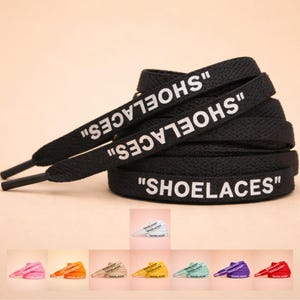 May include: Black shoelaces with the word "SHOELACES" printed in white. The shoelaces are flat and have a woven texture. Additional shoelaces in various colors are displayed below, including pink, orange, beige, yellow, and blue.