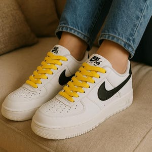 May include: White leather sneakers with black swoosh logos and bright yellow laces. The shoes have a classic design with a low profile and a textured sole. The tongue features the Nike Air logo.