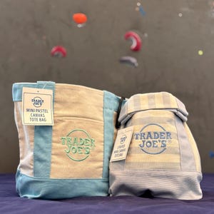 May include: Two canvas tote bags by Trader Joe's. The left bag is cream with light blue trim and features green embroidered text. The right bag has light blue and cream stripes with blue embroidered text. Both bags have attached product tags.
