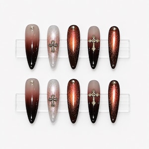May include: A collection of ten long, coffin-shaped false nails. The nails showcase diverse designs, including a brown gradient, silver glitter, and a cross design with rhinestones. Each nail is adorned with a small, round embellishment.