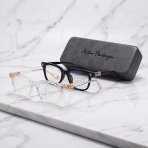 May include: Two pairs of glasses, one with clear frames and one with black frames, both featuring gold-toned metallic accents at the hinges. A grey rectangular glasses case with the text Arthur Pendragon printed in black is positioned behind them.