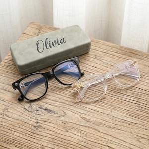 May include: A pair of black-framed glasses and a pair of clear-framed glasses with gold-tone accents on the temples, positioned next to a rectangular grey storage case with the name Olivia printed in black script on the top surface.