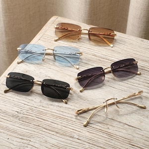 May include: Five pairs of rimless sunglasses with rectangular lenses in brown, light blue, dark gray, and clear tints. Each pair features gold-tone metal temples and bridge with a decorative animal motif on the hinges and arms.
