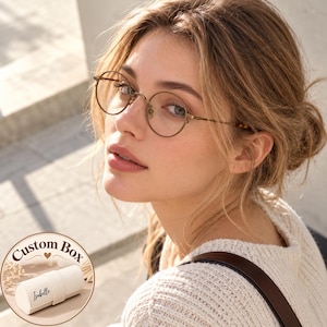 May include: Round-framed glasses with a thin metallic bronze finish. In the corner, a circular inset shows a cream-colored cylindrical jewelry case with the text "Custom Box" and the name "Isabelle" printed in black script on the side.