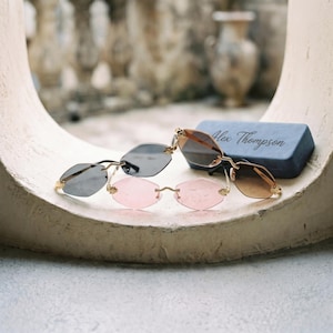 May include: Two pairs of rimless sunglasses with gold-tone metal frames and hexagonal lenses. One pair features dark grey lenses, while the other has light pink lenses. A blue rectangular case with the text Alex Thompson is positioned behind them.
