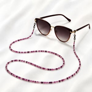 Op de afbeelding: Een zonnebril met een bruin montuur en goudkleurige pootjes, bevestigd aan een lange kralenketting. De ketting bestaat uit kleine, doorschijnende roze en diepmagenta kralen, verbonden aan de bril met zwarte rubberen lusjes.