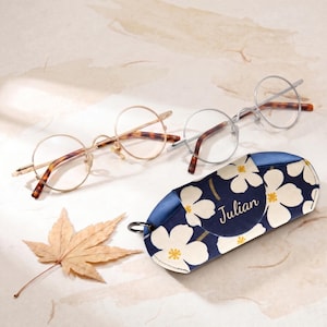 May include: Two pairs of round-frame glasses with thin metal rims and tortoiseshell-patterned temples, one in gold and one in silver, alongside a navy blue fabric glasses case featuring a white floral print and the name Julian in script.