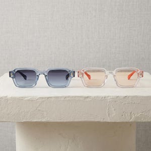 Peut inclure: Deux paires de lunettes de soleil à monture carrée. L'une a un verre dégradé bleu et une monture bleu clair. L'autre a un verre pêche et une monture transparente avec des accents orange. Les deux paires sont exposées sur une surface blanche.