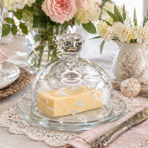 May include: A clear glass butter dish with a domed lid, resting on a decorative plate. The butter is a light yellow color. The dish is surrounded by flowers and a decorative knife.