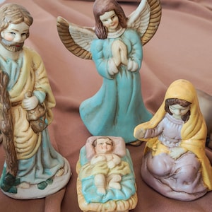 May include: A ceramic nativity set featuring a standing figure with a staff, a winged angel with hands in prayer, a kneeling figure in a yellow head covering, and a small infant lying in a manger, all painted in soft pastel blue, yellow, and tan.