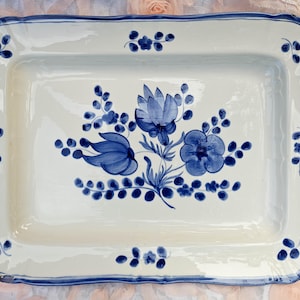 May include: A rectangular ceramic serving platter with a cream-colored base and a hand-painted blue floral motif in the center. The rim features a scalloped edge with a thin blue border and small blue floral sprigs at each corner and side.