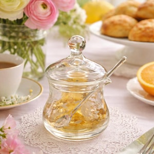May include: A clear glass honey jar with a lid and spoon, set on a white lace doily. The jar is filled with honey and sits on a table with flowers, tea, and pastries.