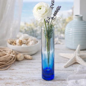 May include: A clear glass vase with a vibrant blue base holds a white flower, lavender sprigs, and delicate white filler flowers. The vase sits on a white wooden surface, with a coastal theme including a starfish and rope.