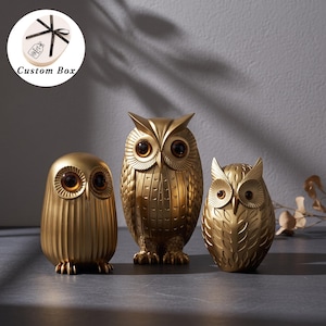 May include: Three gold-colored owl figurines with dark amber eyes. The left owl has vertical ridges, the center owl features detailed feather patterns and tufted ears, and the right owl displays a leaf-like texture. Includes a custom gift box.