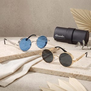 Puede incluir: Dos pares de gafas de sol con lentes redondas y protectores laterales. Un par tiene montura plateada con lentes azules y el otro montura dorada con lentes gris oscuro. Se incluye un estuche cilíndrico negro con el nombre Alexander impreso.