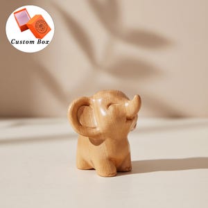 Könnte beinhalten: Eine kleine, glatte, hellbraune Elefantenfigur aus Holz mit rundem Körper, großen Ohren und einem nach oben gebogenen Rüssel. Der Elefant hat einen lächelnden Ausdruck. Eine orangefarbene Geschenkbox mit der Aufschrift Custom Box ist oben links.
