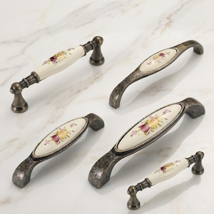 May include: Five cabinet pulls featuring a combination of antique bronze-toned metal and cream-colored ceramic inserts. Each ceramic piece is decorated with a floral pattern of yellow and deep red roses with green leaves and small purple accents.