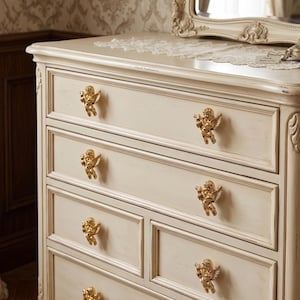 May include: A cream-colored wooden dresser featuring a distressed finish and six drawers. Each drawer is equipped with a polished gold-toned metal knob shaped like a cherub with wings. The dresser has a rectangular top and decorative molding.