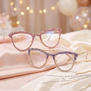 May include: Two pairs of cat-eye glasses with gold-tone frames. The top pair features pink rhinestones and the name Aria printed on the lens. The bottom pair features purple rhinestones and the name Isabelle printed on the lens.