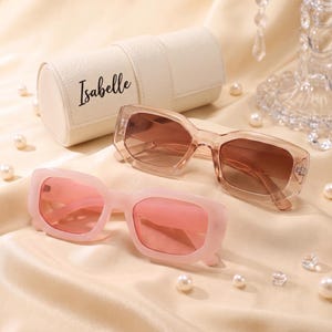 May include: Two pairs of rectangular sunglasses and a cylindrical cream-coloured case with the name Isabelle printed in black script. One pair of sunglasses has a matte pink frame with pink lenses, while the other has a clear frame with brown lenses.