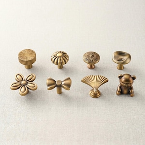 May include: A collection of eight brass-toned metal cabinet knobs in various shapes, including a sunburst, a pumpkin, a floral pattern, a smooth disc, a six-petal flower, a bow, a fan, and a small bear figure, all featuring a metallic finish.