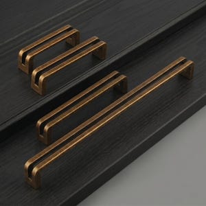 May include: Four metal cabinet pulls with a brushed antique bronze finish. Each handle features a distinct U-shaped profile with a central open channel running the length of the bar. The set includes two shorter pulls and two longer pulls.