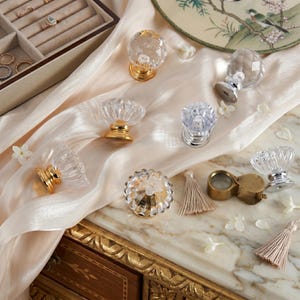 Op de afbeelding: Een collectie van heldere glazen kastknoppen met gefacetteerde oppervlakken en metalen voetjes in goud- en zilverkleuren. De knoppen zijn gerangschikt op een gedrapeerde champagnekleurige stof naast een juweliersloep en twee beige kwastjes.