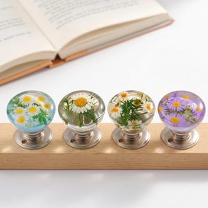 May include: Four round cabinet knobs with clear resin domes containing pressed flowers. The first knob has white daisies on a blue background, the second has a large white daisy, the third has small white daisies, and the fourth has purple flowers.