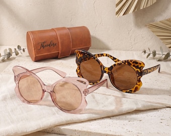 May include: Two pairs of butterfly-shaped sunglasses and a brown cylindrical leather case. One pair of sunglasses is translucent pink with light lenses, and the other is brown tortoiseshell with dark lenses. The case features the name Theodore.