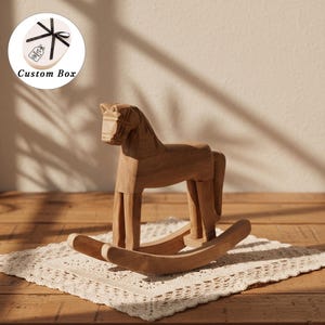 May include: A carved wooden rocking horse with a natural light brown finish and visible wood grain. In the upper left corner, a circular inset shows an oval-shaped gift box with a black ribbon and the text Custom Box printed on it.