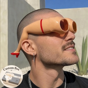 May include: Novelty sunglasses featuring frames shaped like two human legs in a flesh tone, each with a red band above the knee and a small red high-heeled shoe at the temple. The lenses are tinted orange, and the frames are worn on the face.
