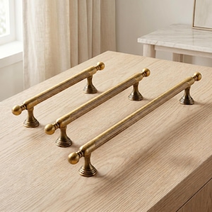 May include: Three antique brass cabinet pulls with a fluted cylindrical bar design, rounded ball ends, and flared pedestal bases. Each handle features a textured, grooved surface along the center section, providing a consistent metallic finish.