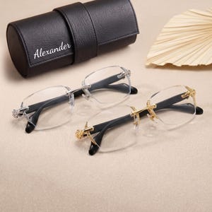 May include: Two pairs of rimless spectacles with black temples, one featuring silver-tone hardware and the other gold-tone hardware, accompanied by a black cylindrical faux leather case with the name Alexander printed in white on the front.