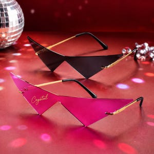 May include: Two pairs of rimless sunglasses with sharp, triangular lenses. One pair features dark black lenses, while the other has vibrant pink lenses with the word Crystal printed in cursive script. Both pairs have thin gold-tone metal arms.