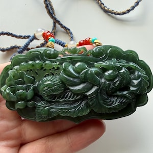 May include: A dark green jade pendant carved with a phoenix and floral motifs, suspended from a braided blue and tan cord. The necklace features decorative accents of red, turquoise, and translucent white beads near the top attachment points.