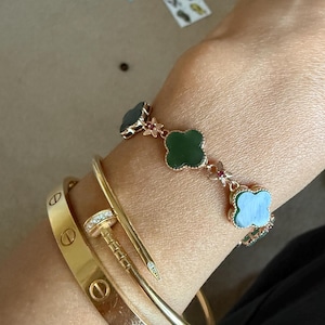 May include: A gold-toned bangle with screw motifs, a gold-toned nail-shaped bracelet with a diamond-encrusted head, and a rose gold-toned chain bracelet featuring dark green clover-shaped charms with small red accents between each clover.
