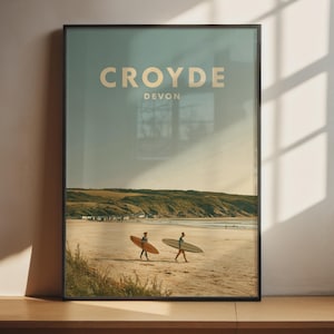Może przedstawiać: Oprawiony wydruk artystyczny przedstawiający plażę, na której dwie osoby niosą deski surfingowe po piasku. U góry znajduje się napis CROYDE DEVON jasnobeżową czcionką. Krajobraz ukazuje piaszczysty brzeg, wzgórza oraz bezchmurne niebo w tle.