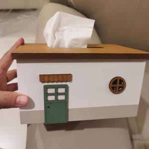 Puede incluir: Caja de pañuelos decorativa con forma de casita. El cuerpo es blanco, con una puerta verde, un tejado marrón y una ventana redonda. Se ven pañuelos blancos saliendo por la parte superior.