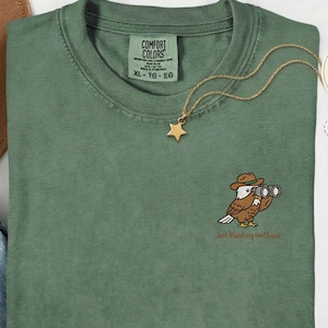 以下が含まれることがあります： モスグリーンのComfort Colors Tシャツ。帽子と双眼鏡を持った漫画のフクロウの刺繍が施されています。「Just Watching the Chaos」の文字が下にあります。金の星のネックレスがシャツの上にあります。