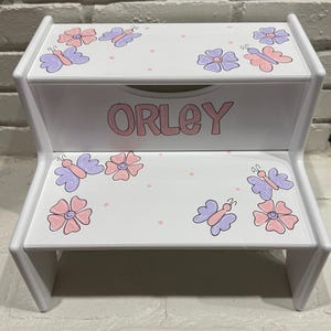 May include: White wooden two-step stool with a pink and purple butterfly and flower design. The top step has the name "ORLEY" painted in pink.