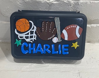 May include: A black lunchbox with a sports theme. The lunchbox has a basketball, baseball glove, baseball bat, football, and baseball. The lunchbox has the name "CHARLIE" written in blue letters.