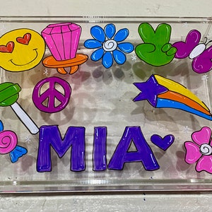 May include: A clear acrylic tray with a colorful design. The tray features a peace sign, a lollipop, a candy, a ring, a flower, a shooting star, a butterfly, a smiley face, and the name MIA written in purple.