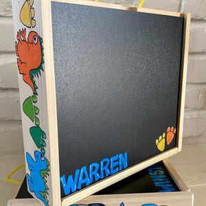 May include: Wooden easel with a black chalkboard surface. The easel has a yellow rope handle and is decorated with dinosaur and rocket ship illustrations. The name "WARREN" is written in blue letters on the chalkboard.