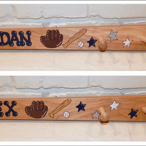 May include: Wooden baseball bat-shaped coat rack with two personalised names, "JORDAN" and "ALEX", painted in navy blue. The rack features a baseball glove, bat, and ball painted on it, along with white and grey stars. There are five wooden hooks on the rack.