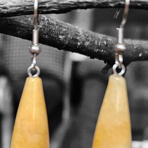 Op de afbeelding: Een paar druppelvormige oorbellen met een lichtgele tint. De oorbellen hangen aan goudkleurige haken en steken af tegen een vage, grijstinten achtergrond. De oorbellen hangen aan een donkere tak.