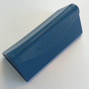 May include: A teal glasses case with a textured finish. The case features a curved top and a snap closure. The words "Plein Les Mirettes" are embossed on the front.