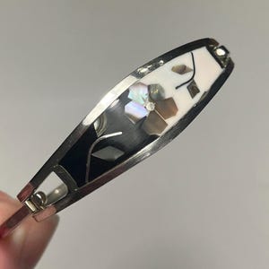 Peut inclure: Bracelet articulé argenté avec un motif floral. Le bracelet présente une incrustation noire et blanche avec des accents de coquillage irisé. Le motif floral est posé sur un fond noir, avec des pétales blancs et irisés.